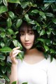 A woman standing in front of a bush of green leaves.