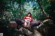 A woman in a red riding hoodie sitting on a rock in the woods.